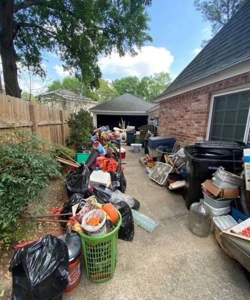 About Coast Junk Removal: A cluttered driveway filled with various discarded items including bags, boxes, and household goods, leading to a garage.