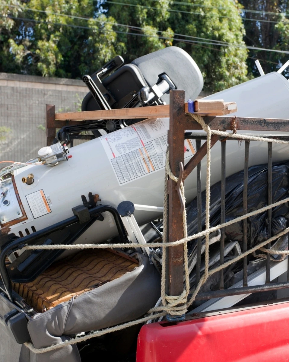 A commercial services pickup truck bed filled with various items including a wheelchair, a water heater, and pieces of wood, all secured with rope.