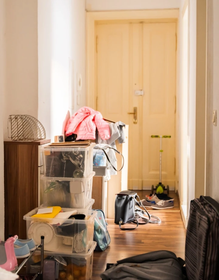 A cluttered hallway in a commercial services building, lined with stacked transparent storage bins, bags, shoes, and a scooter, leading to a closed door.